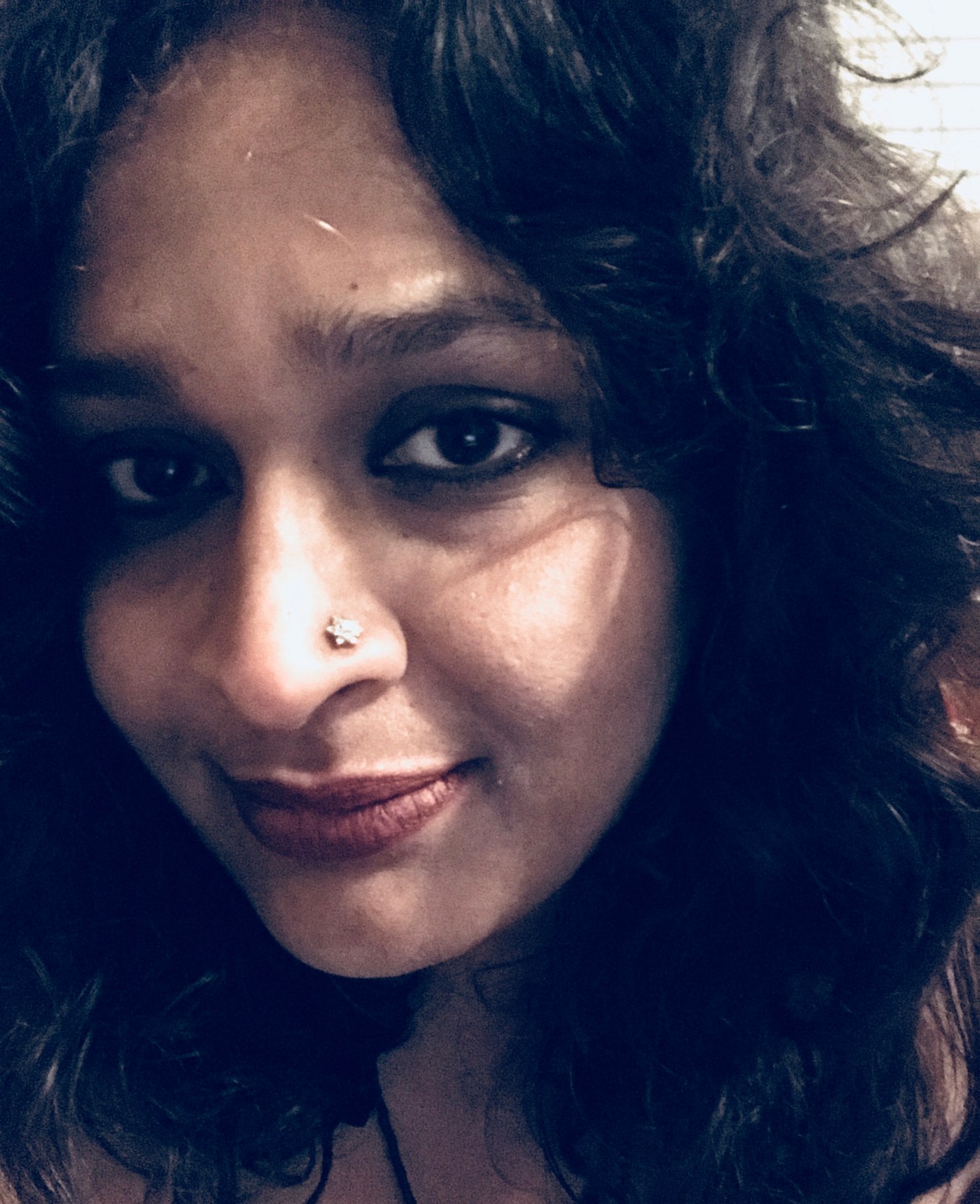 A color photo showing a selfie of an unsmiling Indian woman, with curly dark brown hair and dark eyes. She has a nose stud on her left nostril.