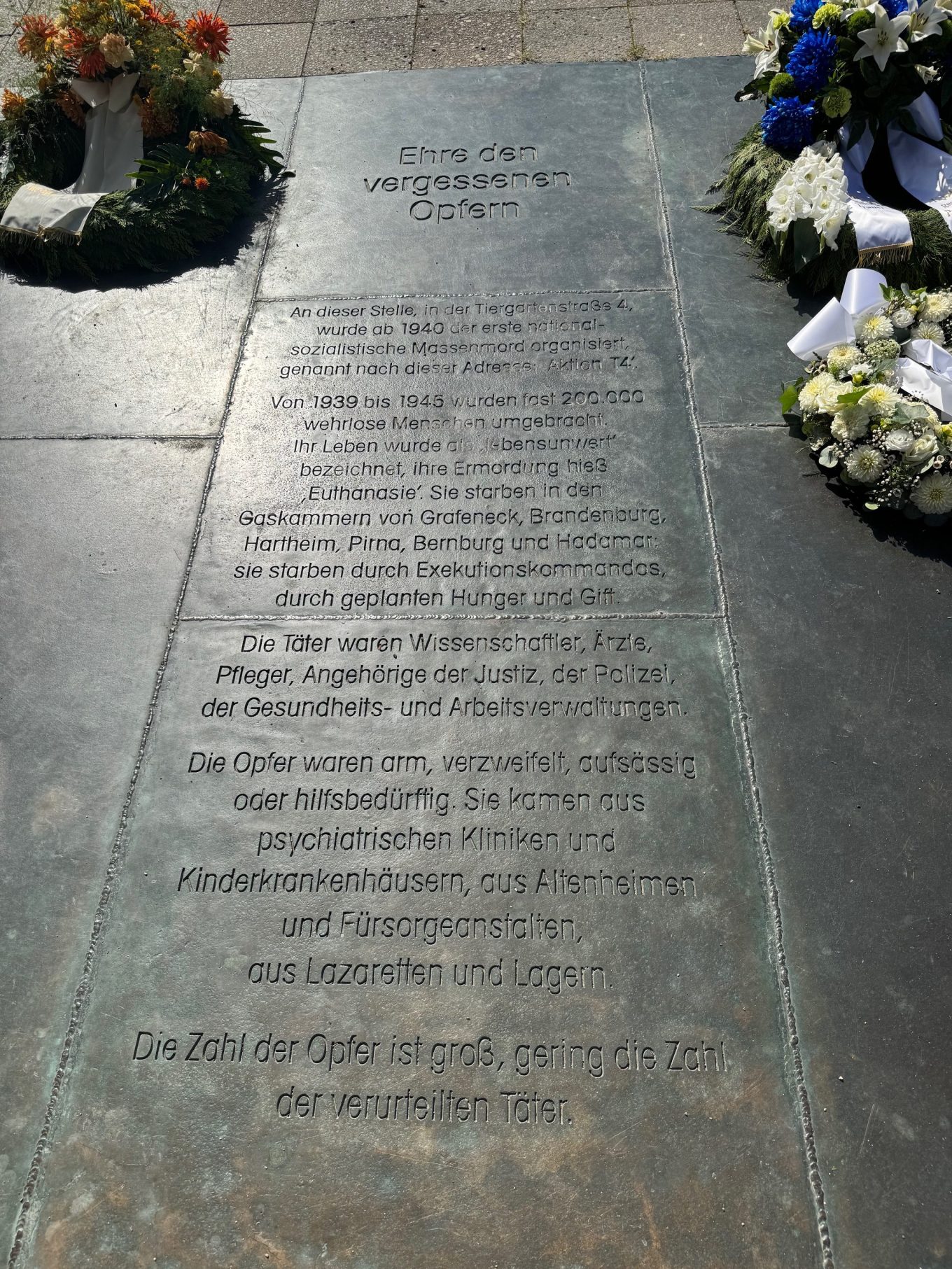 Photo of a memorial plaque at Tiergartenstraße 4 in Berlin. The inscription reads: “Honor to the forgotten victims. At this site, at Tiergartenstraße 4, the first National Socialist mass murder was organized starting in 1940, named after this address: Aktion T4. From 1939 to 1945, almost 200,000 defenseless people were killed. Their lives were described as ‘unworthy of life,’ their murder called euthanasia. They died in the gas chambers of Grafeneck, Brandenburg, Hartheim, Pirna, Bernburg, and Hadamar; they died by firing squads, by deliberate starvation, and by poison. The perpetrators were scientists, doctors, nurses, members of the judiciary, the police, and of the health and labor administrations. The victims were poor, desperate, defiant, or in need of help. They came from psychiatric clinics and children’s hospitals, from nursing homes and welfare institutions, from military hospitals and camps. The number of victims was vast; the number of perpetrators convicted was very small.”