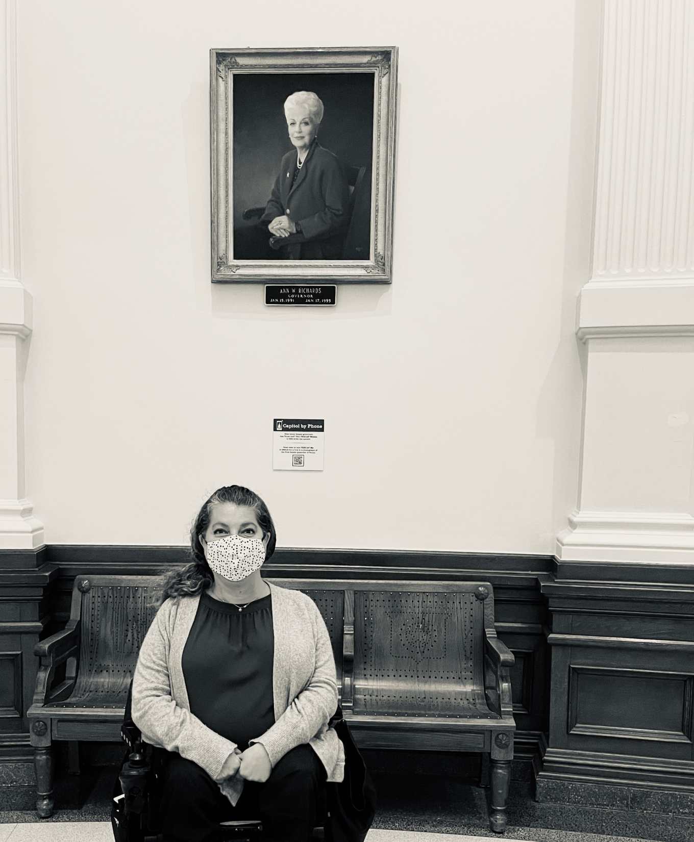 A black and white picture of Emily Wolinsky sitting in her power wheelchair in front of a portrait of the late Governor of Texas, Ann Richards. 