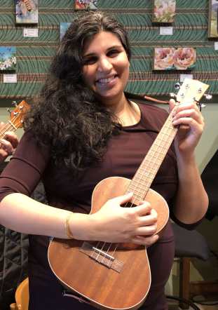 A brown woman with long, curly, black hair, stands smiling while playing a ukulele. She is visibly pregnant, wearing a dark purple dress. The background shows a green wall with small paintings of flowers hanging.