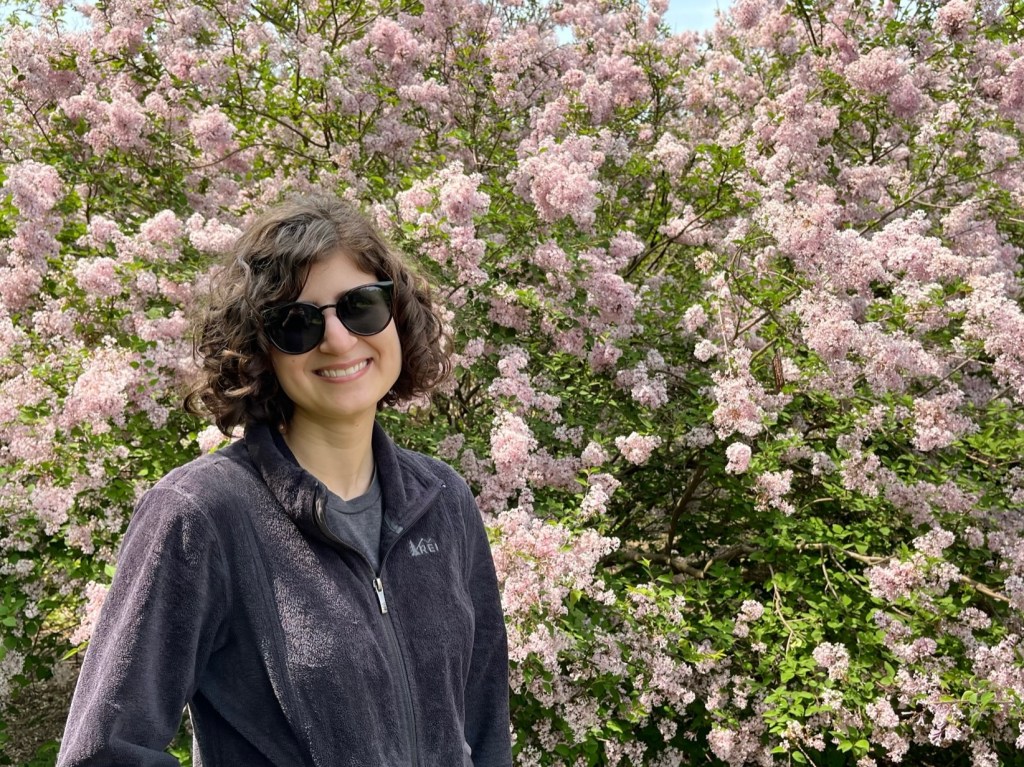 A photo of Raina, a smiling white woman with short curly brown hair. She is wearing sunglasses and a black jacket and is in front of a flowering lilac tree.