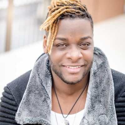 Picture of a Black man with blonde dreadlocks smiling at camera wearing a black ribbed sweater jacket with grey fur collar, white t-shirt and two silver necklaces. Photo credit: RJ Patel