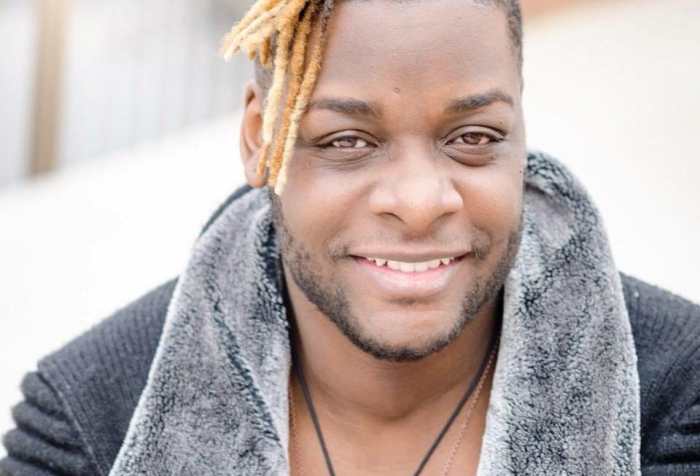 Picture of a Black man with blonde dreadlocks smiling at camera wearing a black ribbed sweater jacket with grey fur collar, white t-shirt and two silver necklaces. Photo credit: RJ Patel