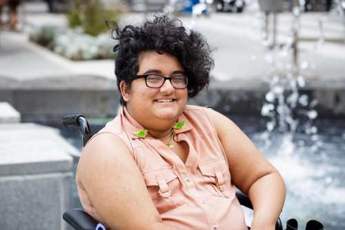 Noor sits in a wheelchair in front of a fountain. He has a curly black undercut and medium golden tan skin. He wears thick glasses, and a sleeveless pink shirt with a set of budgie collar pins.