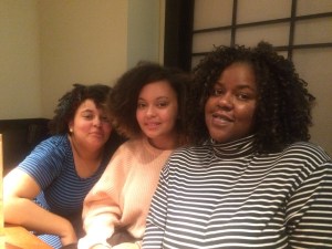 A Black woman with curly hair wearing a striped turtleneck. The the left of her are her two teenage biracial daughters, both with curly hair.