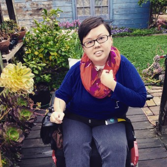 Image description: photo of Stacey in her garden. She has short hair, is mixed race Asian American, and uses a power wheelchair. She is wearing a bright orange scarf and is surrounded by succulents.