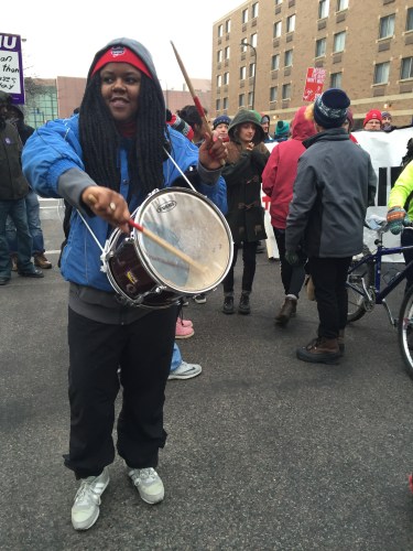 A Black woman with long braids wearing a blue jacket with a hoodie. She has a drum around her body and is holding two drumsticks.