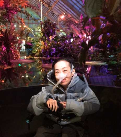 Photo on the of an Asian American disabled woman in a wheelchair in front of a pool of water with tropical water plants in the background lit with purple & magenta lights inside a greenhouse. She is wearing a puffy gray jacket and a mask over her nose attached to a tube.