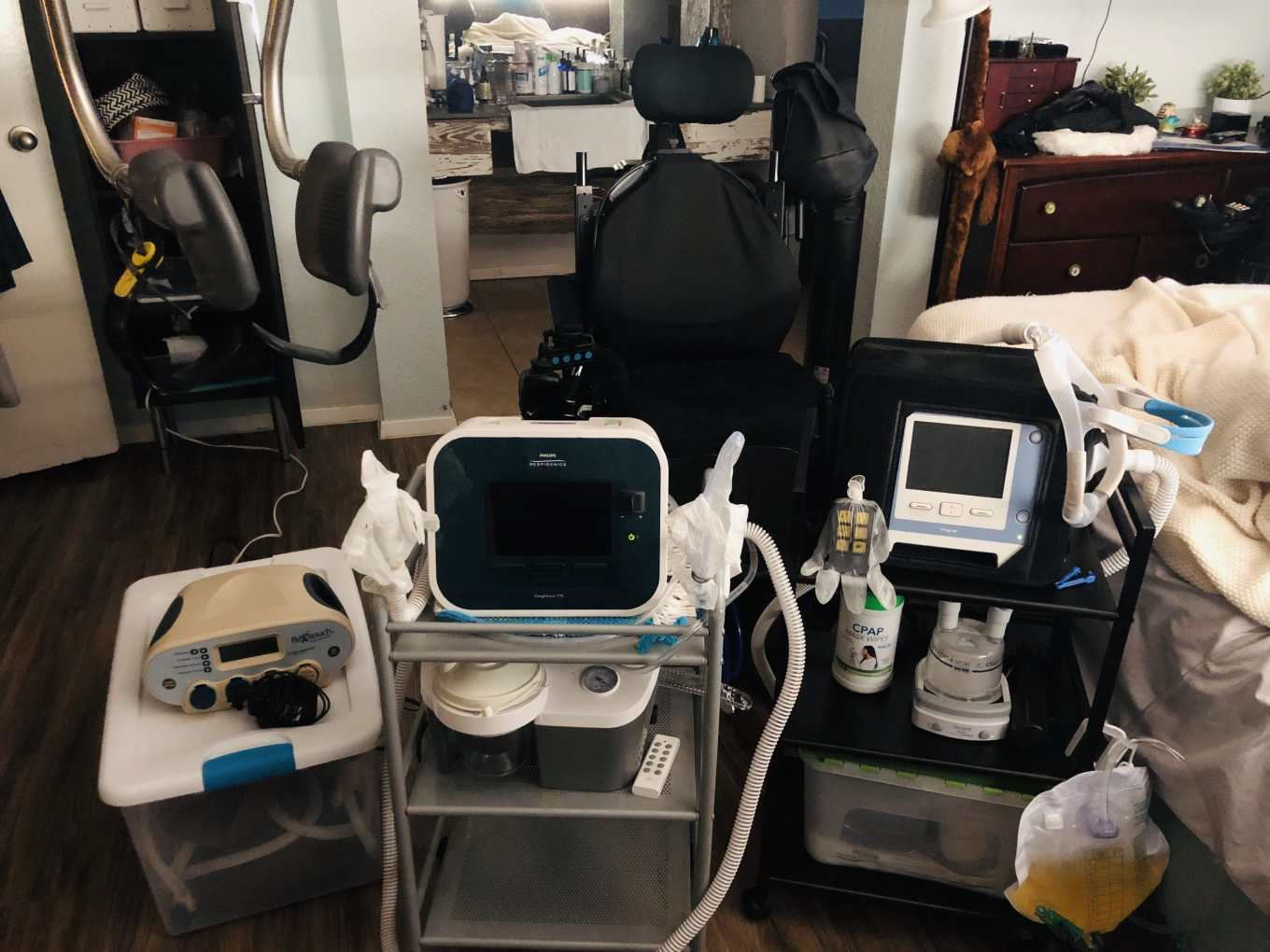 Emily’s durable medical equipment. Top Left: My SureHands ceiling lift named "J-Lo." Bottom Left: My lymphedema therapy machine named "Jose." Middle Front: My Cough Assist named "Sir Coughs Alot" and underneath it, my suction machine, "Sir Sucks Alot." Back Front: Power Wheelchair named "Tina Turner" Left: My Trilogy vent and humidifier named "Darth" sit on a three-tiered cart. J-Lo's remote is also placed on the cart, covered in a latex glove to prevent it from getting wet.   Hooked to the Bottom of the Cart: A disgusting bag of warm, yellow, body waste named "Ted Cruz" that's attached to my suprapubic catheter, "PeeTee" [not pictured].