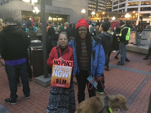Two women of color at an outdoor protest. One is holding a sign that says, "No Peace. Justice Now! BLM" The other woman is holding the leash with a dog