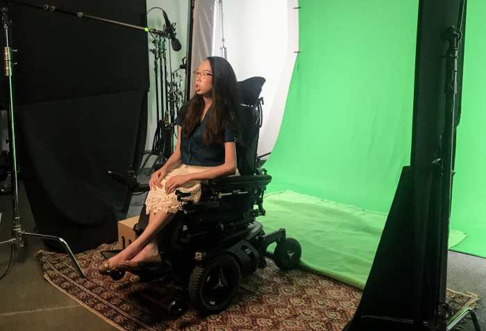 Aubrie on a film set in front of a green screen and with a boom mic pointed at her. She wears a teal blouse, white lace skirt, and black glasses. She rides a power wheelchair.