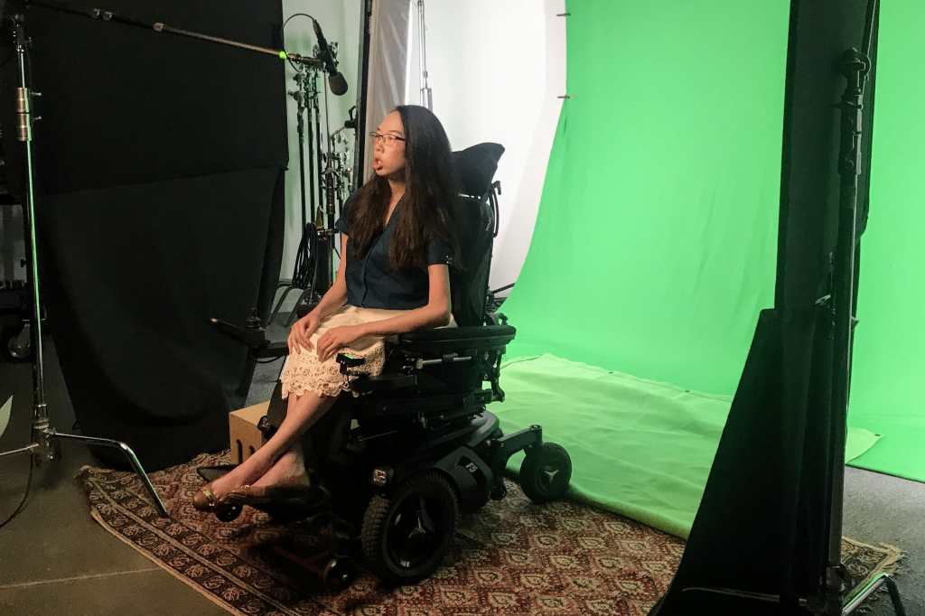 Aubrie on a film set in front of a green screen and with a boom mic pointed at her. She wears a teal blouse, white lace skirt, and black glasses. She rides a power wheelchair.