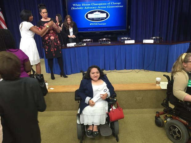 Image of Sandy Ho, a young Asian American woman in a wheelchair in front of a stage at the White House. Behind her is a long table with a blue tablecloth and a large television screen showing the logo of the White House.