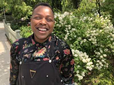 Cyrée Jarelle Johnson, a black transmasculine person with medium brown skin, a septum ring, and a big smile stands in front of a hydrangea bush that is behind a white picket fence on a sunny day. It’s so bright his eyes have squinted closed. He is wearing black shortalls, a gold triangle necklace, and a silky, floral black shirt rolled up to the elbows. Photo by J.D. Stokely