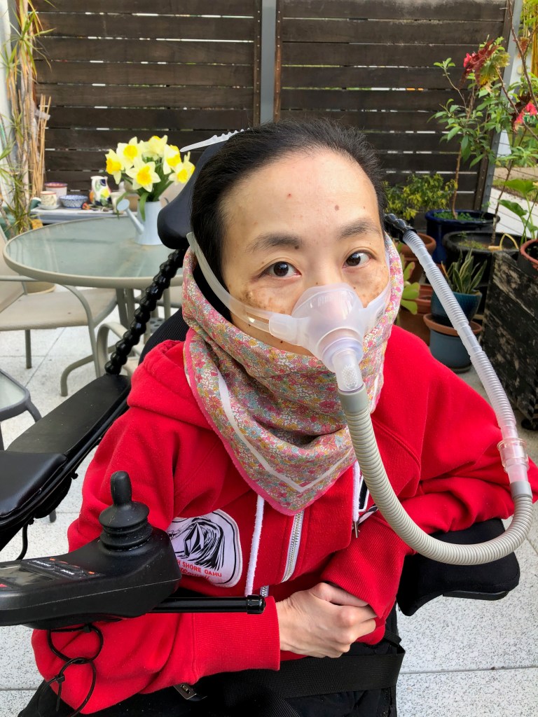 Asian American woman in a power chair sitting at a patio with a red hoodie and a floral print handkerchief around her mouth. She’s wearing a mask over her nose attached to a gray tube.