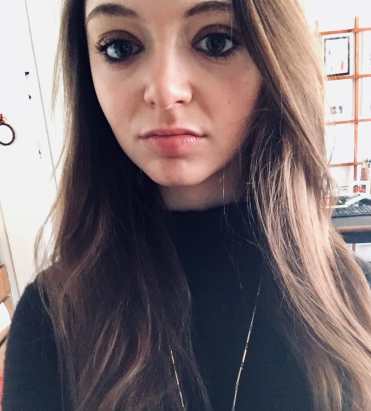 A woman with long wavy brown hair stands, smiling. Her face fills almost the entire photo frame. She has brown-colored eyes, brown eyeshadow, and pink lipstick while wearing a long sleeve black turtleneck shirt. Behind her is an office wall covered with small artworks and photographs.