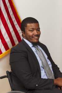 Image of a black man smiling with arms crossed in front of flag