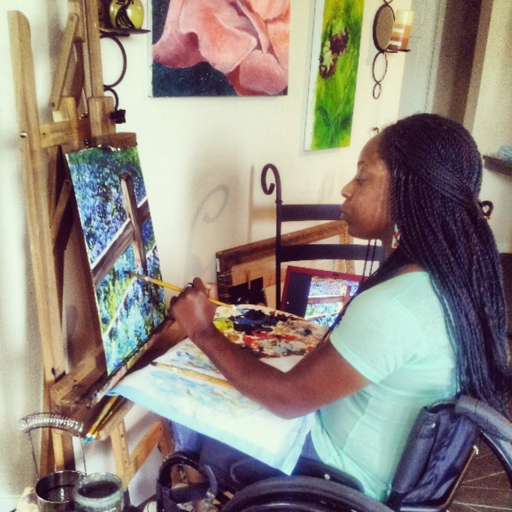 Photo of Kemi Yemi-Ese, a young Nigerian American Black woman with long black braids. Kemi is painting at her easel while sitting in her manual wheelchair. The canvas is of a wooden fence and a field of bright blue-purple flowers.