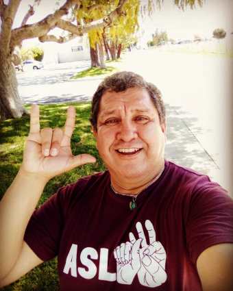 Photo of Hector M. Ramirez, a Latino Chiricahua Apache man wearing a maroon t-shirt that says ASL with 3 illustrations of hands spelling the letters a-s-l. His right hand is showing the sign, "I love you" in American Sign Language.