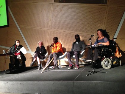 Image of a stage with five people sitting and having a conversation. There is an Asian American woman in a wheelchair on the very left with a small table in front of her. To her right is a white woman with white-blonde hair and a black dress. Next to her is an older Black disabled man with a bright orange shirt and a wooden walking stick. Next to him is a younger Black man with long dreadlocks and a beard. He is wearing a gray top and dark pants. The final person on the right side of the stage is a woman of color in a wheelchair with a blue top and a table connected to her wheelchair.