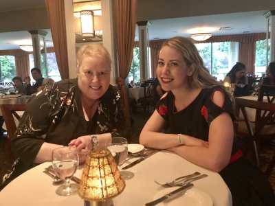 Two women with blonde hair smiling and posing for the camera at a table at a fancy restaurant in Harrison Hot Springs. On the left is Cathleen wearing a dark flower printed blouse and on the right is Amanda, the writer, wearing a black dress with roses on the sleeves. They look happy and rested.