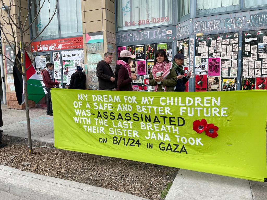 Photo of Samar AlKhdour wearing a light pink baseball cap and a red and white Keffiyeh standing in conversation with a sit-in supporter in front of Canadian Minister of Immigration Marc Miller’s riding office in Montreal on April 29, 2024. Behind them are the Immigration minister’s outside office-building walls covered in posters and stickers in support of Palestinian liberation. In the foreground, there is a bright green banner with the words: “My dream for my children of a safe and better future was assassinated with the last breath their sister Jana took on 8/1/24 in Gaza.” The lettering is white and black. There are also red Palestinian poppy flowers sewn into the banner. Photo by Nelly Bassily