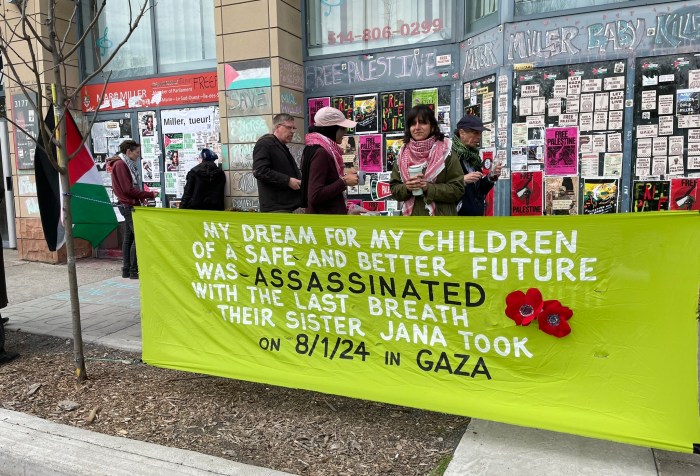 Photo of Samar AlKhdour wearing a light pink baseball cap and a red and white Keffiyeh standing in conversation with a sit-in supporter in front of Canadian Minister of Immigration Marc Miller’s riding office in Montreal on April 29, 2024. Behind them are the Immigration minister’s outside office-building walls covered in posters and stickers in support of Palestinian liberation. In the foreground, there is a bright green banner with the words: “My dream for my children of a safe and better future was assassinated with the last breath their sister Jana took on 8/1/24 in Gaza.” The lettering is white and black. There are also red Palestinian poppy flowers sewn into the banner. Photo by Nelly Bassily