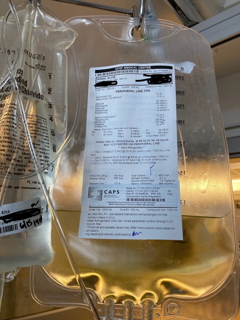 A large plastic bag filled partially with a yellow fluid of nutrition to be delivered intravenously. A label lists the contents and date prepared. On the left is another plastic bag connected to tubing with a clear liquid.