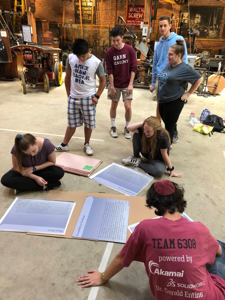 A group of students looking at posters for a museum exhibit that are laying on the ground. Photo courtesy of Gann Academy. Photo credit: Maya Wainhaus
