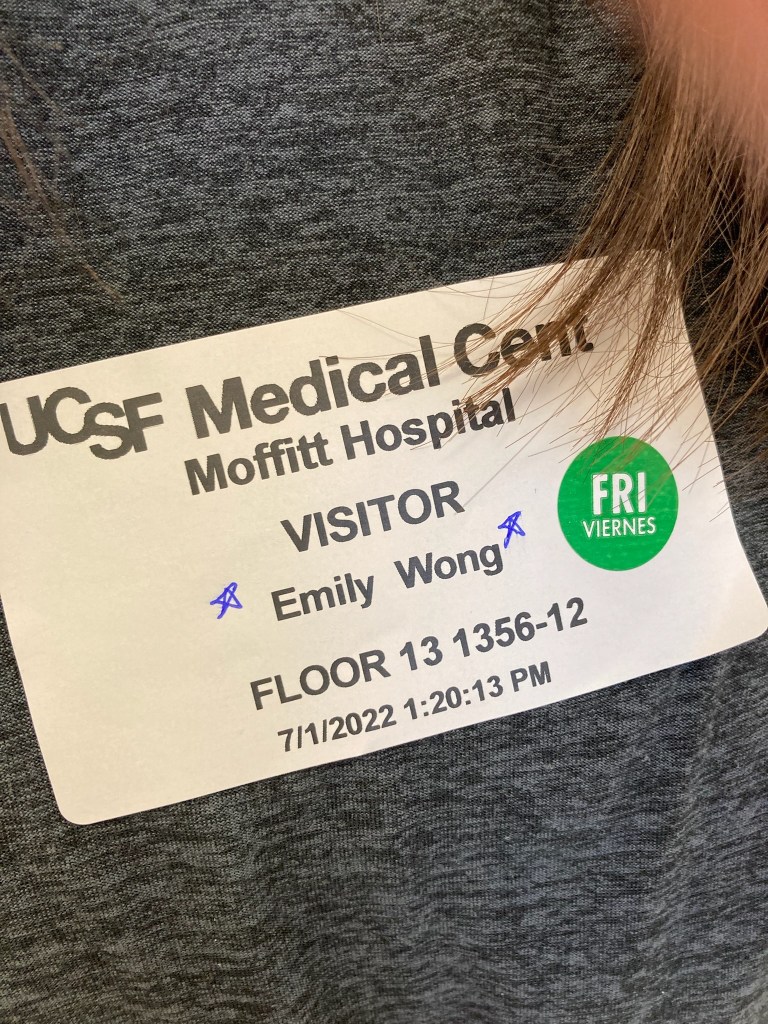 A visitor’s badge on my sister’s gray shirt with the text: “UCSF Medical Center Moffitt Hospital VISITOR Emily Wong Floor 13, 1356-12, 7/1/2022, 1:20:13 PM.” The stars on the badge indicate she is allowed to stay overnight.