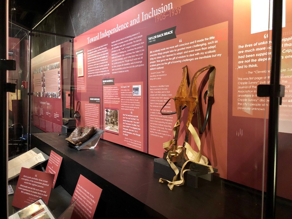 Disability history exhibit featuring a back brace, a baseball glove, and an early hearing aid. The title for this section: "Towards Inclusion and Independence 1905-1939." Photo courtesy of Gann Academy. Photo credit: Maya Wainhaus