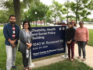 Outdoor photo with the PRIDE project team in front of a campus sign that reads: "Disability, Health and Social Policy Building, 1640 W. Roosevelt Rd., Heart Center & Physical Therapy" there are large trees in the background.