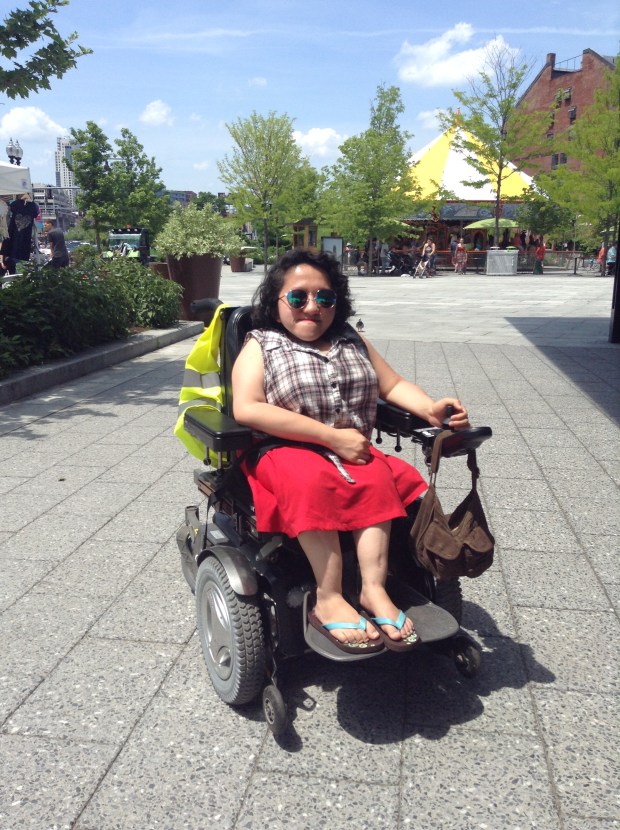 Image of Sandy Ho, a young Asian American woman in a wheelchair, outdoors on a sunny day. She is wearing sunglasses.
