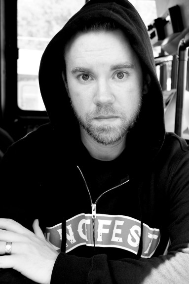 Black and white photo of writer Adam Pottle, a young white man wearing a black hoodie. He is staring intently at the camera. His arms are crossed with each hand resting on the opposite arm.