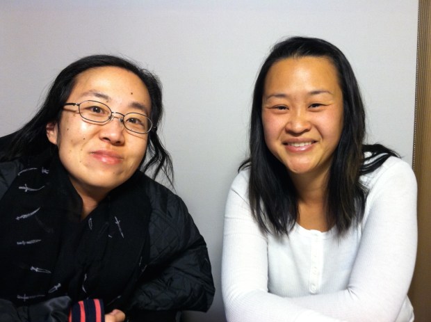 Two Asian American women sitting side-by-side smiling at the camera. The woman on the right is wearing a black jacket and glasses. The woman on the right is wearing a white shirt and has long black hair.