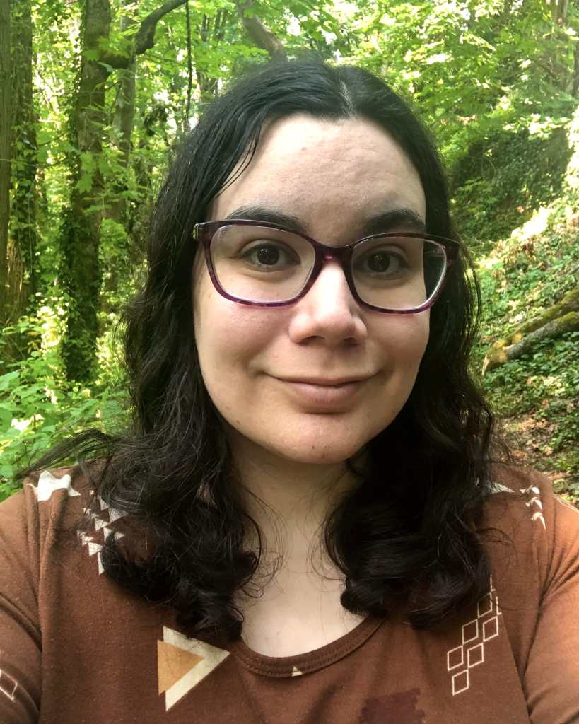 A headshot of a woman with light brown skin and shoulder-length black, wavy hair wearing purple glasses and a brown and orange patterned shirt. She’s smiling. There’s a forest in the background.