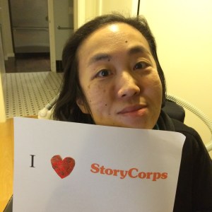 Image of an Asian American woman in a wheelchair holding a paper sign that reads, "I [heart symbol] StoryCorps"