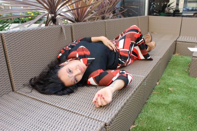 Photo of a young woman with long dark hair lying on a outdoor patio furniture in the shape of a couch, one hand is resting on her stomach while the other is lying against the surface of the couch. She has a dress that has an abstract print in black, red-orange and white. The person featured in this image is a self described, 'a black, gross femme, queer punk writer, dancer/actor/model with arthrogryposis'