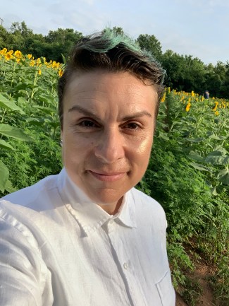 L. Harris, a white, androgynous person with brown and green short hair and wearing a white shirt, with a field of sunflowers behind them.