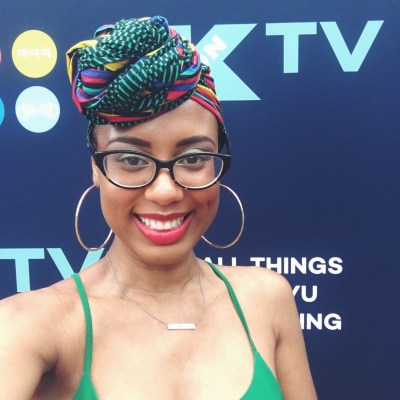 Tiffany smiles radiantly with dimpled cheeks wearing large gold hoop earrings and a colorful Haitian headwrap with swirls of bright green, yellow, black and red. Her black & brown tortoise shell glasses rest atop her nose. 