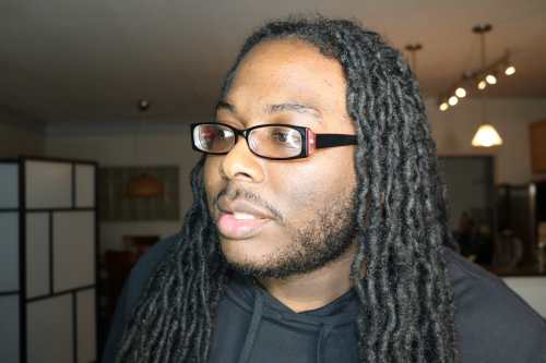 Da'Shaun is standing in a room with his head turned to the left glaring at something off camera. His hair is hanging down, he has on red and black glasses, and his mouth is opened slightly—a partial grin, of sorts