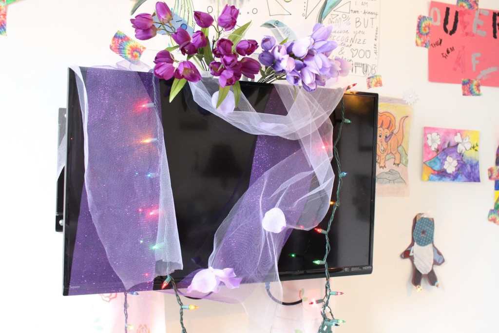 [The television in Emet’s hospice room. There is purple and white tulle, multicolored rainbow lights, and purple flowers draped over it. In the background art is visible on the walls.]