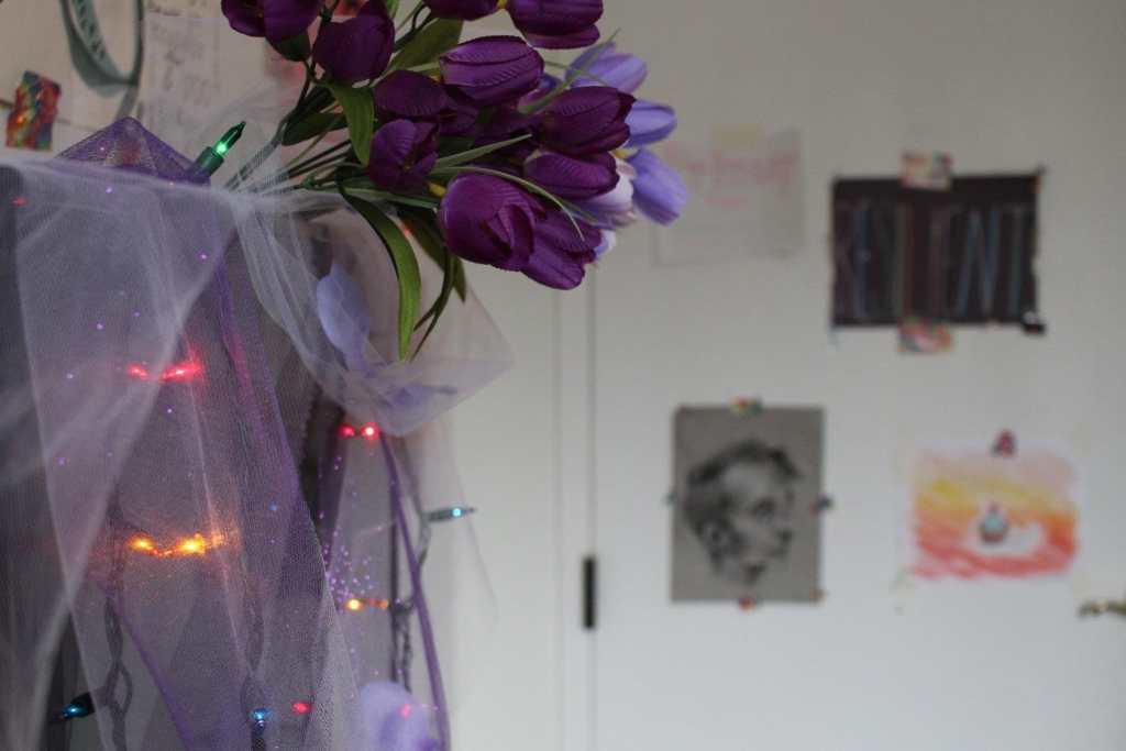 [A profile shot of the television in Emet’s hospice room. Purple tulle, multicolored lights, and purple flowers are draped over it. In the background the door is visible, covered in pieces of art made by queer and trans people.]
