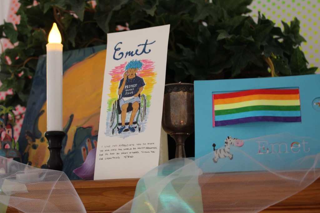 [A blue card with a rainbow flag, a zebra sticker, and the name Emet. A white paper card with Emet in black cursive writing. Under it is a rainbow background with a drawing of Emet, a white disabled trans man with bright blue hair, sitting in his wheelchair, wearing a black t-shirt and gray shorts. His t-shirt says “Protect Trans Kids” in white letters. He is holding a speech in his lap. The card is signed by Nico. A white candle is next to the card. White tulle is draped over the dresser, as well as blue ribbon. A plant is in the background.]
