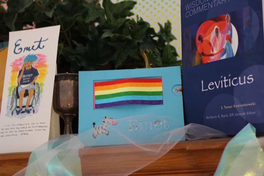 [A blue card with a rainbow flag, a zebra sticker, and the name Emet. A white paper card with Emet in black cursive writing. Under it is a rainbow background with a drawing of Emet, a white disabled trans man with bright blue hair, sitting in his wheelchair, wearing a black t-shirt and gray shorts. His t-shirt says “Protect Trans Kids” in white letters. He is holding a speech in his lap. A white candle is next to the card. White tulle is draped over the dresser, as well as blue ribbon. A plant is in the background. On the right is a dark navy blue copy of Leviticus.]