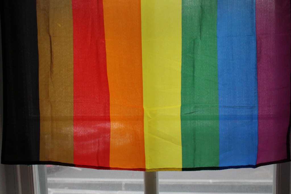 [A more color more pride progress rainbow pride flag, draped over a window.]