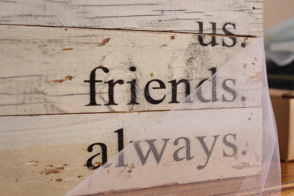 [A white wood block with the words “us. friends. always.” in black letters. It is draped in white tulle.]