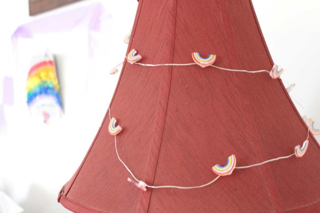 [A red lampshade with rainbow multicolored string lights draped over it. In the distance is a rainbow, with purple tulle draped over it.]