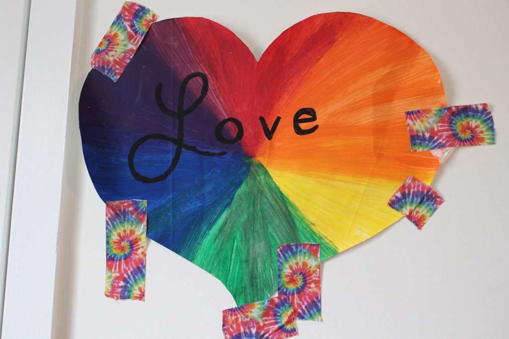 [A rainbow heart with the word Love in large black lettering. It is attached to the wall with rainbow tie dye tape.]