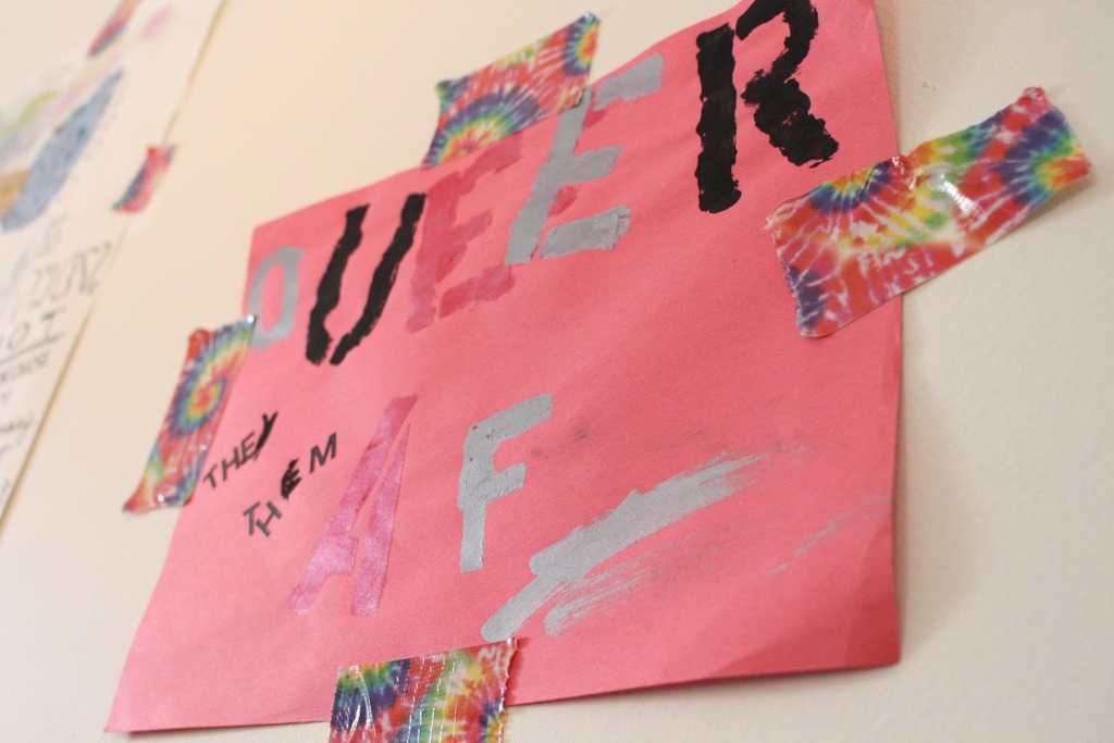 [“Queer AF” in alternating Red, Silver, Black paint, on red paper. “THEY THEM” in black handwriting. It is attached to the wall with rainbow tie dye tape.]